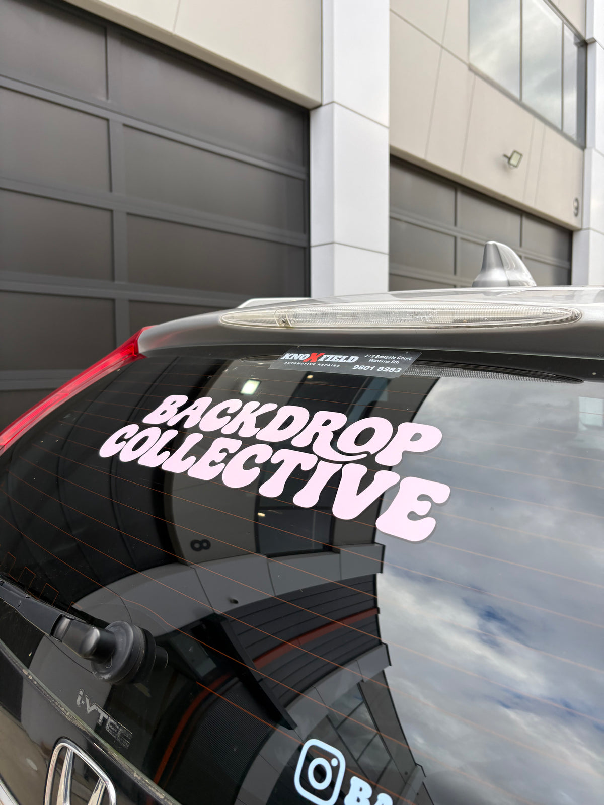 A vinyl decal sticker reading 'BACKDROP COLLECTIVE' in bubbly white letters with a pink shadow, applied to the rear window of a black car, shown outdoors in front of a garage door.